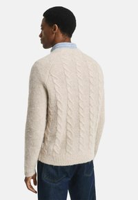 Man in een crème kabelgebreide trui over een blauwe kolshirt en donkere spijkerbroek, met de rug naar ons gekeerd tegen een witte achtergrond.