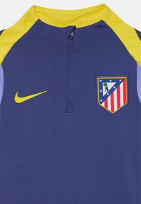 Nike trening majica u plavoj boji s žutim detaljima, zatvaranje na pola patentnog zatvarača i izvezeni logotip Atlético Madrida s prugama i zvijezdama.