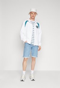 Chaqueta ligera blanca con acentos en verde azulado, camiseta a rayas, pantalones cortos de mezclilla, sombrero de cubo blanco y zapatillas en tonos neutros.