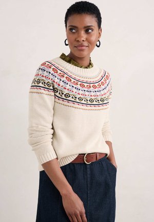 Jersey de punto color crema con patrones geométricos coloridos a lo largo del pecho, puños y dobladillo acanalados; combinado con una blusa verde con cuello y una falda de mezclilla oscura.