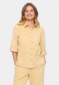 Light yellow button-up shirt with a collar, roll-up sleeves, and brown buttons. Soft fabric with a relaxed fit, paired with matching pants.