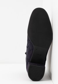 Semelle de chaussure habillée en daim foncé avec semelle noire, inclinée contre un mur blanc, montrant le nom de la marque "högl" sur la zone de la voûte plantaire.