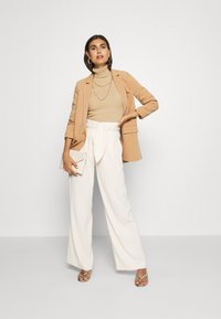 Beige blazer over a beige ribbed turtleneck, white high-waisted wide-leg trousers, holding a light clutch, with gold jewelry and strappy sandals.