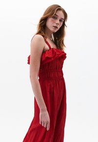 Rød flæsedragt med en smocked talje, tynde stropper og et flydende design med vertikal tekstur. Stoffet ser let og luftigt ud.