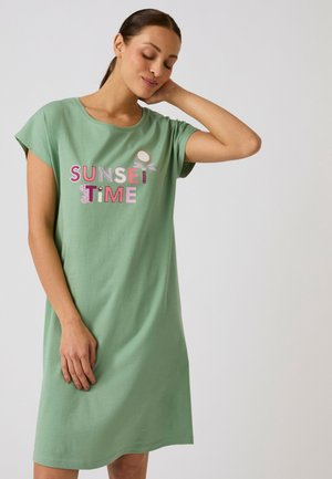Vrouw in een groene knie-lange jurk met "SUNSET TIME" tekst in roze en wit, staand met één hand achter haar hoofd, ogen gesloten.