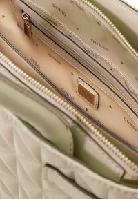 Sac à main beige matelassé avec un compartiment zippé, présentant un intérieur lisse avec des logos « GUESS » répétés et de petits motifs triangulaires.