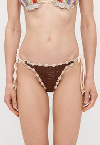 Bruine gehaakte bikini onderkant met gekartelde randen, voorzien van beige strikjes met houten kralen aan beide zijkanten. Geweven look, minimale bedekking.