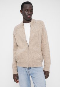 Beežist lukuga kinnitatav kardigan tekstuurse kudumiga, kõrge krae ja soonikuga mansettidega. Esiosas taskud ja peened triibud kogu pinnal.