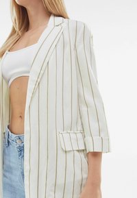 Bílý pruhovaný blejzr se zlatými vertikálními pruhy, s jednou náprsní kapsou a ohrnutými rukávy. Nositý přes bílý crop top, v kombinaci se světle modrými džíny.