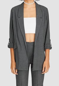 Blazer gris à fines rayures avec une coupe décontractée, col à revers crantés et manches retroussées. Associé à un crop top blanc et un pantalon assorti.