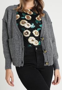 Szary sweter kardigan z ribbowanymi mankietami, z trzema dużymi guzikami, przednimi kieszeniami i luźnym krojem, nałożony na top w kwiatowy wzór.