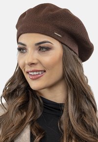Bruine gebreide baret met een gestructureerde afwerking, voorzien van een kleine metalen accent. Het zit comfortabel op het hoofd met een zachte, slungelige draping.