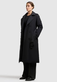 Zwarte trenchcoat met een dubbelgebreid ontwerp, grote kraag en schouderbanden. Gemaakt van een gladde stof met een slanke, moderne silhouet.