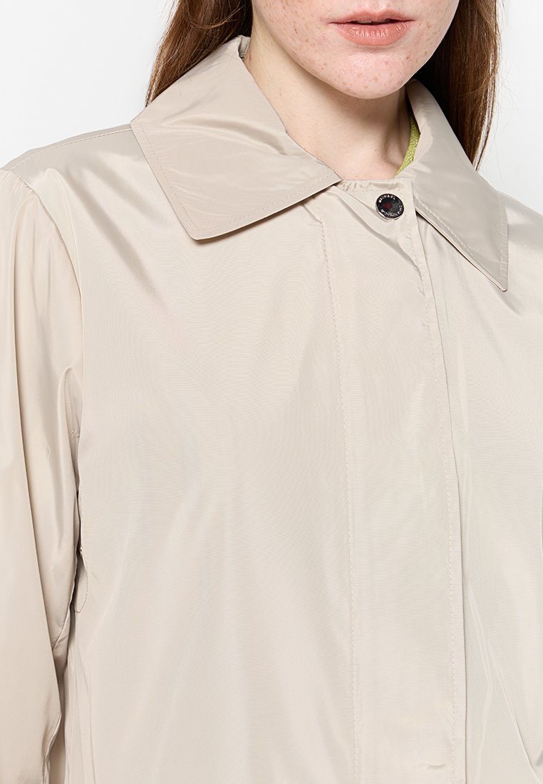 Veste beige légère avec un grand col pointu, fermeture à bouton-pression et éclat subtil ; elle comporte des poches latérales et une coupe décontractée.
