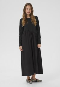 Fekete, hosszú ujjú ruha, összegyűrt derékkal és lenge szoknyával; fekete-fehér csíkos balerina cipővel párosítva. Egyszerű, modern dizájn.