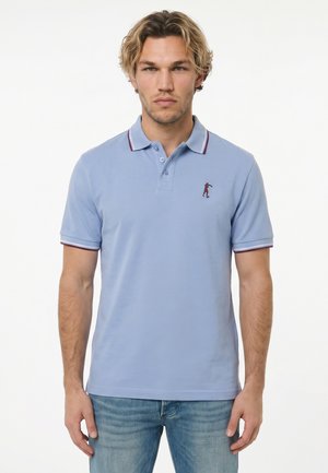 Uomo con capelli biondi mossi che indossa una polo azzurro chiaro con colletto a righe bordeaux e bianco e jeans azzurri chiari.