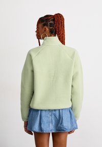 Femme aux cheveux tressés rouges portant de grandes boucles d'oreilles, une veste en polaire vert clair et une jupe en denim bleu courte de dos.