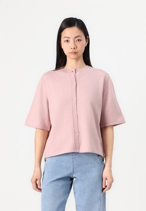 Cardigan rose clair à manches courtes avec un col rond, en tissu texturé et fermeture par boutons devant. Associé à un jean en denim bleu.