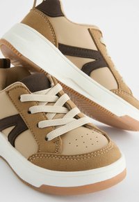Baskets beige et brunes en cuir et daim, semelle en caoutchouc blanche, à lacets. Comprend des perforations sur le bout.