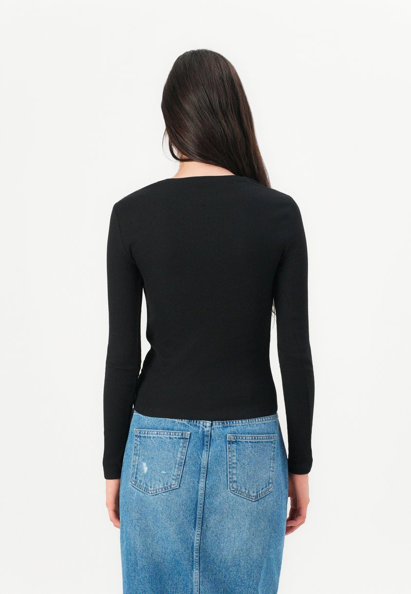 Kobieta z długimi ciemnymi włosami, ubrana w czarną bluzkę z długim rękawem oraz niebieską dżinsową spódnicę, odwrócona plecami na jednolitym białym tle.