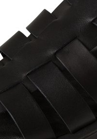 Black leather sandal featuring wide interwoven straps and a textured surface. The design has a flat sole and minimal stitching details.