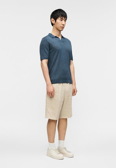 Marineblå poloskjorte laget av et mykt stoff, kombinert med beige linshorts. Modellen har på seg hvite joggesko og sokker, og står foran en hvit bakgrunn.