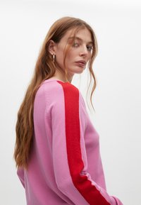 Rosa Pullover mit langen Ärmeln, der einen auffälligen roten Streifen auf der linken Schulter hat. Glatte Textur, klassisches Rundhalsdesign.