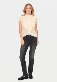 Kortom, een lichtgele blouse met korte mouwen en een gerimpelde kraag en voorkant, met een subtiel patroon, gecombineerd met donkere jeans en zwarte schoenen.