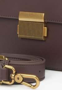 Fialová kožená taška s klopou ve zlatém odstínu a s vertikálními drážkami. Obsahuje odpovídající popruh se zlatým otočným háčkem.