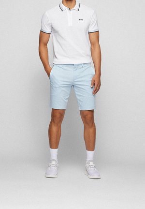 Witte poloshirt met zwarte randen, gecombineerd met lichtblauwe shorts. Model draagt witte sneakers met witte sokken en staat in een neutrale pose.