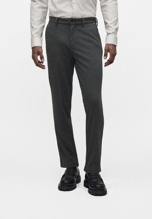 Muž oblečený do tmavosivých slim-fit nohavíc, čiernych masívnych kožených topánok a svetlosivej košele s dlhým rukávom, stojaci s uvoľnenými rukami.