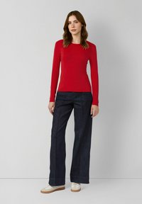 Czerwona przylegająca bluzka z długim rękawem zestawiona z szerokimi, ciemnoniebieskimi dżinsami. Modelka ma na sobie białe sneakersy z beżowymi akcentami. Prosty design, gładka tekstura.