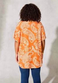 Blouse orange à motif cachemire blanc, coupe ample, manches courtes et encolure ronde. Associée à un jean bleu ajusté.