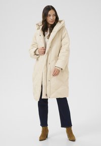 Beige puffebluser med hætte, der har et quiltet design og trykknaplukning. Bæres over en lys skjorte med knapper og mørke jeans.