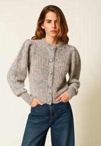 Giovane donna con capelli castano chiaro sciolti che indossa un cardigan grigio lavorato a maglia con grandi bottoni e jeans blu scuro a gamba larga, con le mani nelle tasche.