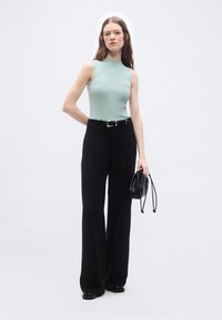 Γυναίκα που φοράει αμάνικη ανοιχτοπράσινη μπλούζα, μαύρο παντελόνι με φαρδιά μπατζάκια και μαύρη ζώνη, μαύρα παπούτσια, κρατώντας μικρή μαύρη τσάντα, σε απλό φόντο.