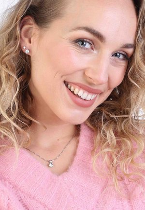Zilveren ketting met een hanger, vergezeld van studs. Het model heeft golvend, schouderlang haar en draagt een zachtroze trui.