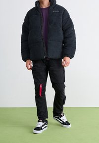 Černá fleecová bunda s vysokým límcem, zapínáním na zip a nadýchaným designem. Nahoře fialové tričko, černé cargo kalhoty a černé tenisky.