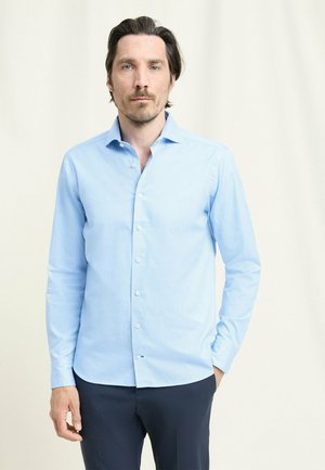 Man met donker haar, gekleed in een lichtblauw overhemd met knopen en een donkere broek, staand tegen een eenvoudige lichte achtergrond.