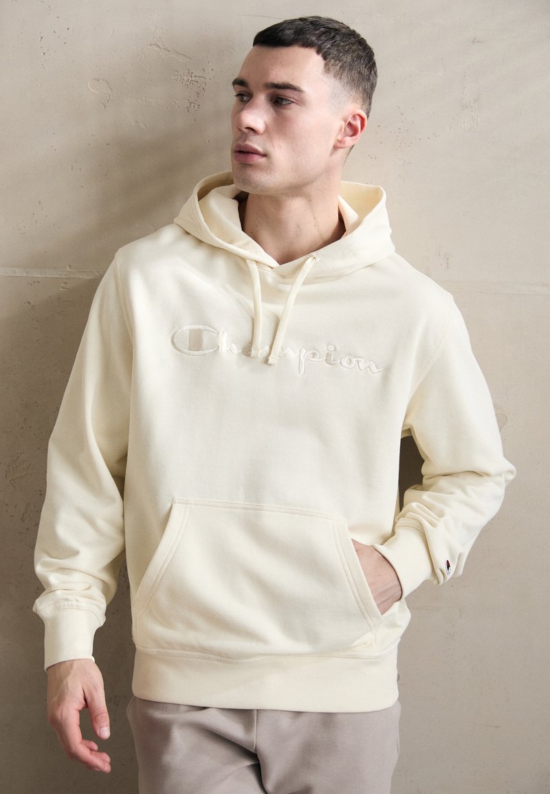 Krémový mikina vyrobená z mäkkého materiálu, s predným vreckom, sťahovacou kapucňou a vyšitým logom "Champion" na hrudi.