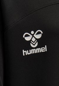 Svart atletisk tyg med en vit hummel-logotyp, som visar två vingliknande former ovanför en stiliserad chevrondesign. Texturerad yta är synlig.