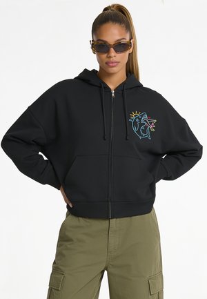 Femme avec queue de cheval portant des lunettes de soleil rectangulaires marron, un sweat à capuche noir zippé avec un motif coloré de requin et un pantalon cargo vert olive.