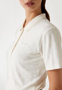 Cremefarbendes Poloshirt mit strukturiertem Muster und dezentem, wiederholtem Logo-Design. Kurze Ärmel und ein klassischer Kragen mit gestreiften Akzenten.