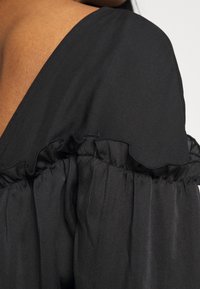 Primer plano de una prenda negra de volantes sin hombros llevada sobre un hombro de tono de piel, con detalles de tela recogida.