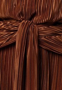 Glänzender bronzefarbener, plissierter Stoff, der gerafft und zu einem großen Knoten gebunden ist und dabei Textur und reflektierenden Glanz hervorhebt.