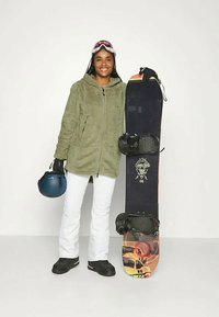 Grüne Fleecejacke, weiße Hose, schwarze Stiefel; hält ein Snowboard mit grafischem Design, schwarzen Bindungen und einem blauen Helm. Skibrille auf dem Kopf.