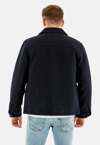 Mørkeblå fleecejakke med en skarp krave, lange ærmer og knapmanchetter, parret med lyseblå jeans, som fremviser et simpelt design.