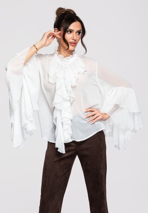 Femme aux cheveux foncés attachés en chignon, portant un chemisier blanc transparent à volants et un pantalon marron foncé, posant avec une main sur la hanche et l'autre près du visage.