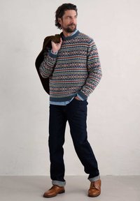 Strickpullover mit blauen, roten und cremefarbenen geometrischen Mustern, kombiniert mit dunklen Jeans und braunen Lederschuhen, mit einer braunen Jacke lässig über die Schulter gelegt.