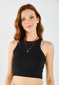 Top corto negro de canalé con un escote alto. Se lleva con dos collares en capas: uno de cuentas rojas y una cadena dorada con un colgante de corazón.